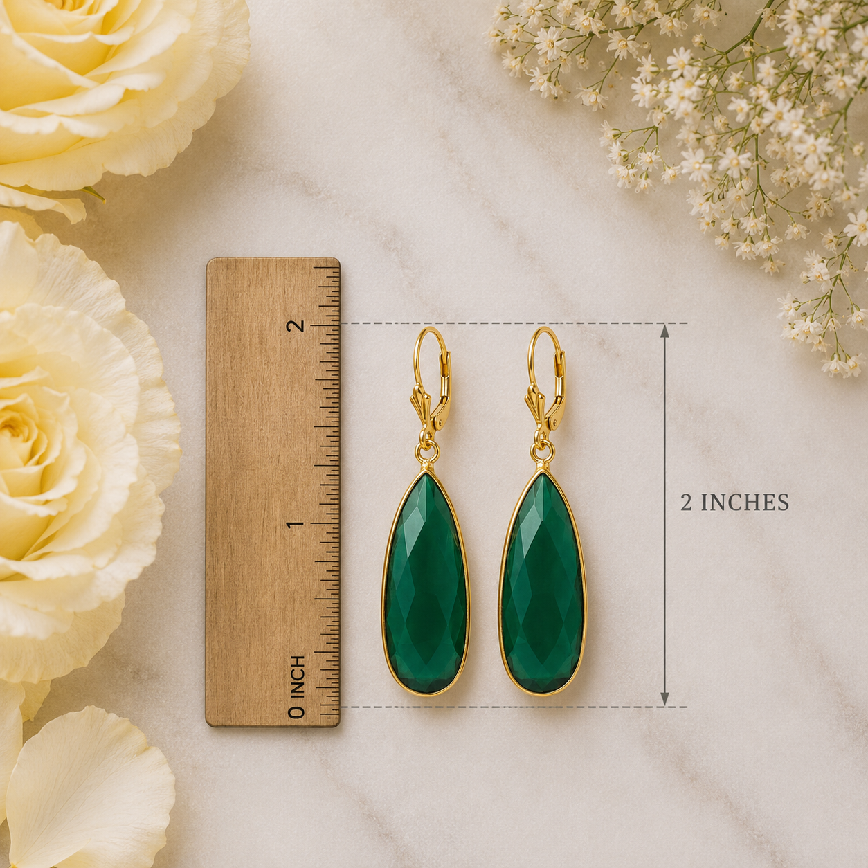 Green teardrop earrings with gold accents on a marble surface with flowers and a ruler for scale.